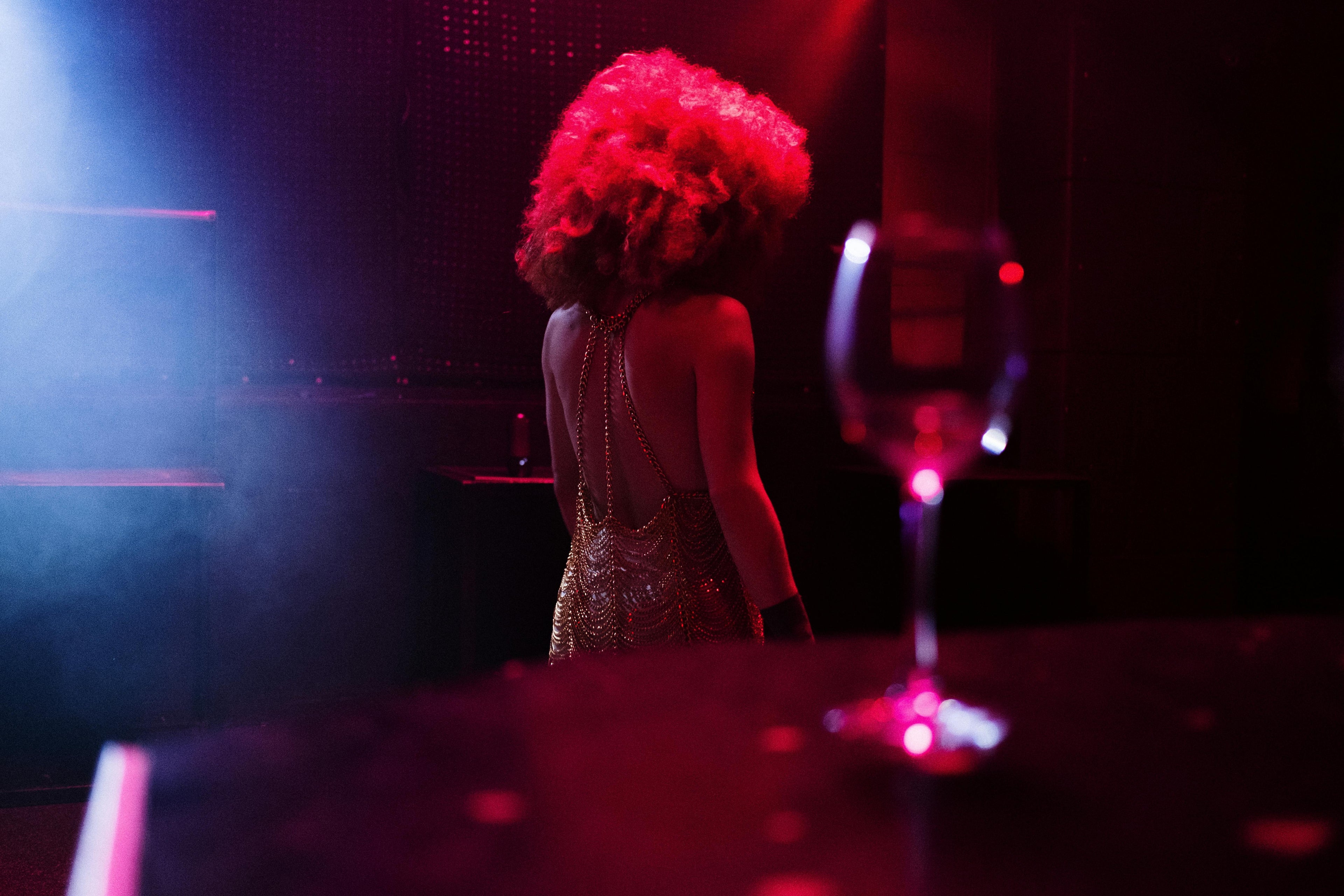 Personne vue de dos dans une ambiance de club aux lumières rouges et violettes, portant une robe dorée scintillante.